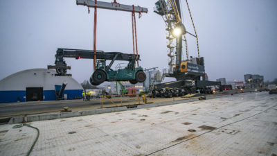 Reachstacker hängt am Haken des Hafenmobilkrans mit Schwerlast-Traverse. Im Hintergrund Lagerhallen