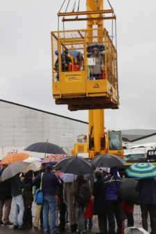 Menschen mit Regenschirmen vor einem gelben Autokran, der einen Aussichtskorb in die Höhe zieht